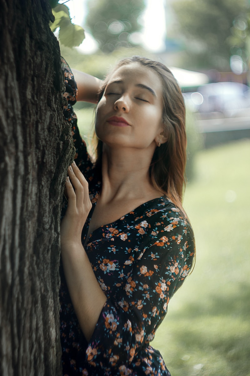 woman in black and green floral spaghetti strap top leaning on brown tree trunk during daytime